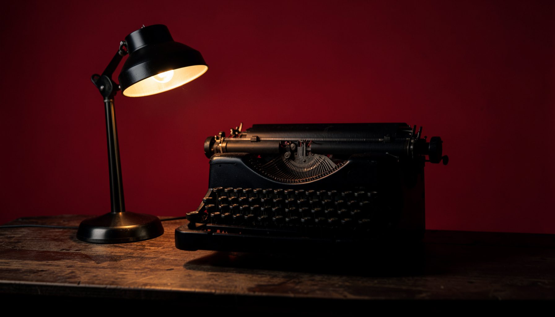 Dark atmospheric photograph of a vintage typewriter on a wooden desk, lit by a single warm lamp, deep charcoal tones with subtle amber highlights